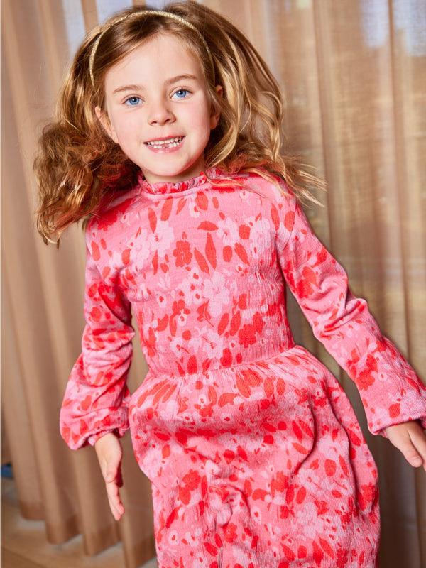 A child wearing the Izzy eco-friendly kids smock dress in pink, pictured mid-jump and looking into the camera.