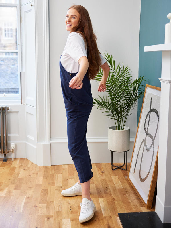 A model wearing the Kathleen dungarees in navy, pictured playfully swinging their arms  in front of a potted plant and a painting.