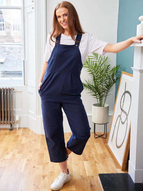 A model wearing the Kathleen dungarees in navy, pictured in a bright room in front of a fireplace. 