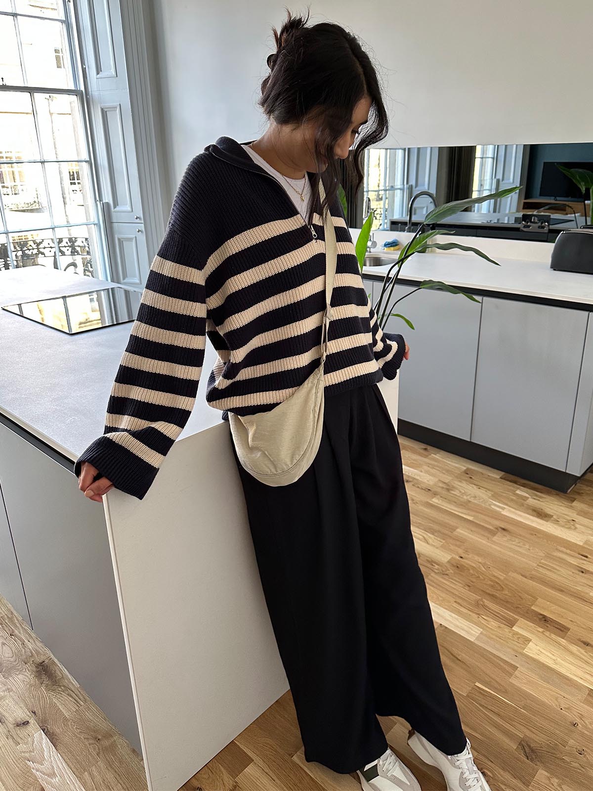 Model wearing the Lana sustainable trousers in black with a striped black and white jumper and a white cross body bag. They are in a well lit room, leaning against a countertop with their face mostly hidden by their hair as they look towards the ground. 