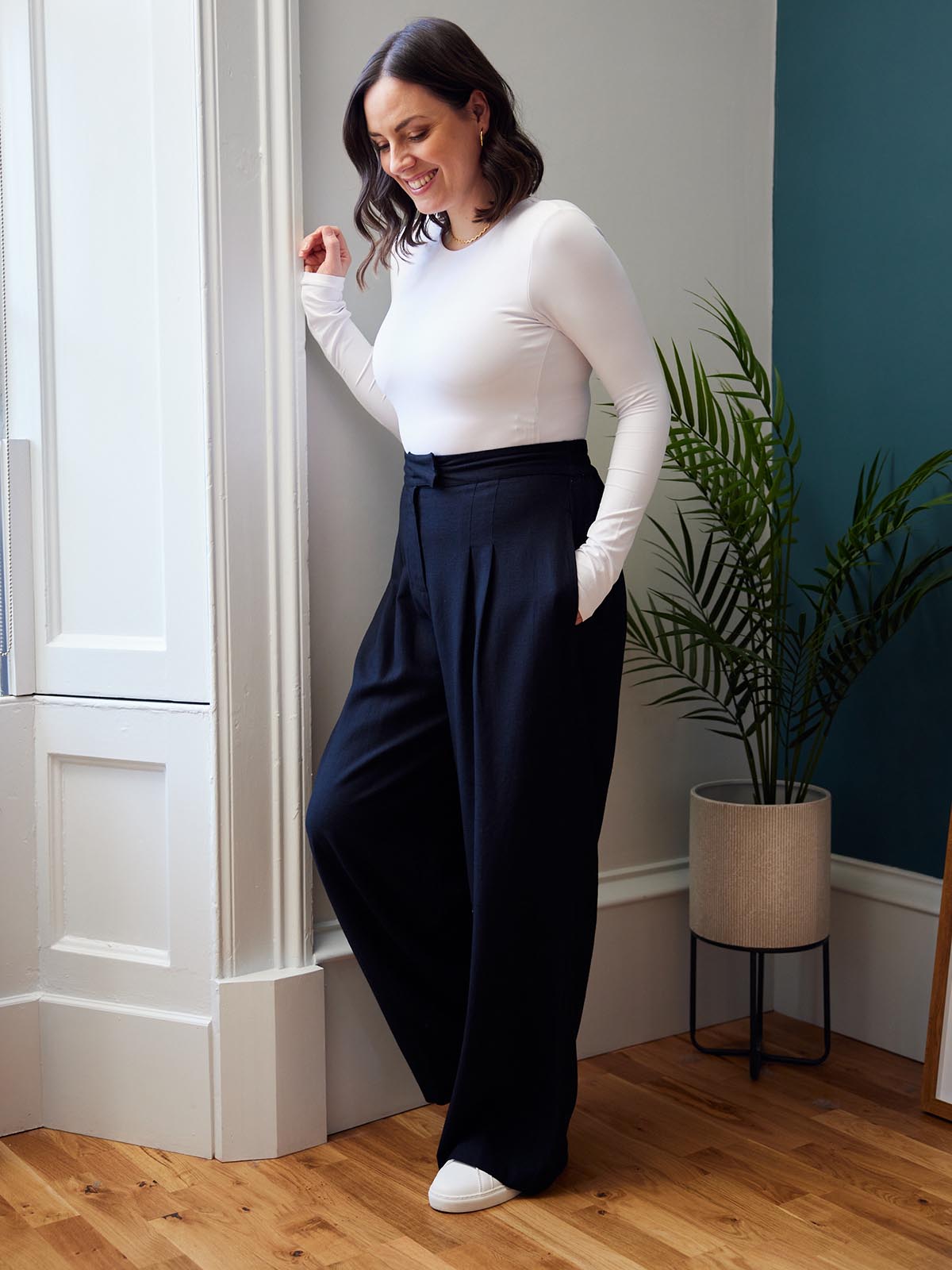 Model wearing the Lana sustainable trousers in black and a long sleeve white t-shirt, pictured leaning against a wall and smiling at the ground in a living room in front of a potted plant.