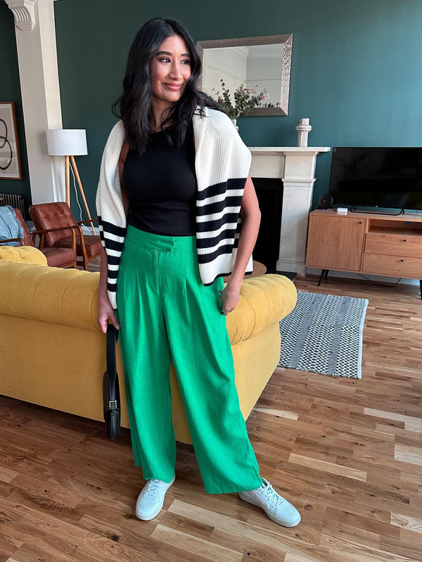 A model wearing the green Lana trousers, trainers and a black vest top pictured in a living room holding a handbag and smiling 