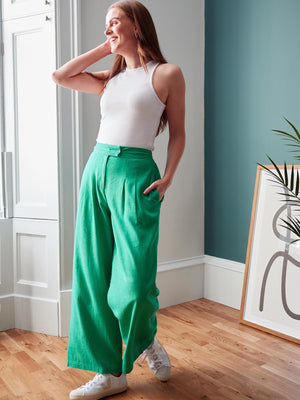 Model wearing the green Lana trousers and a white vest top, pictured looking to the side and brushing their hair back with their hand.