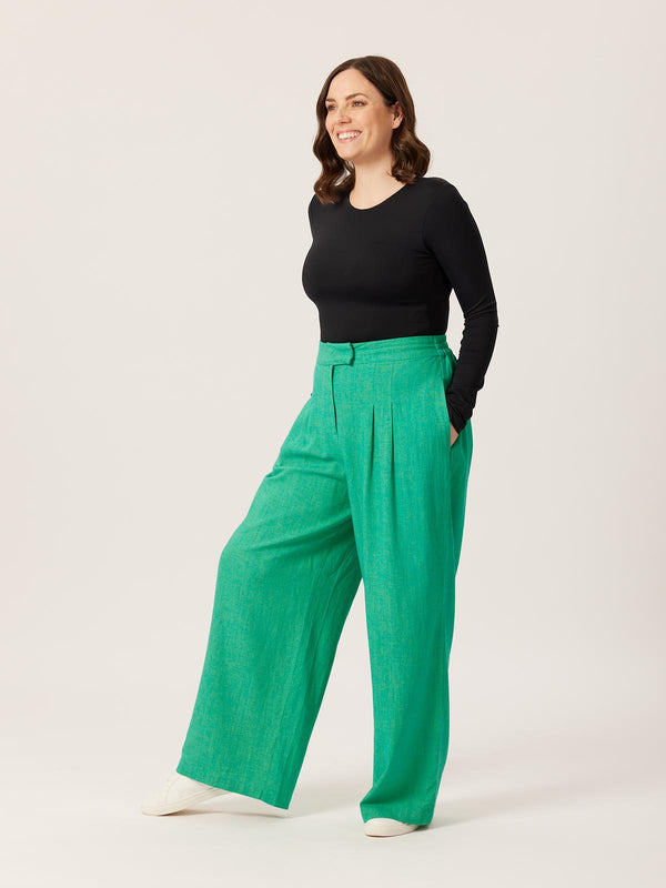 A model wears the green Lana trousers with a long sleeve black t-shirt, pictured with their hands in their pockets. 