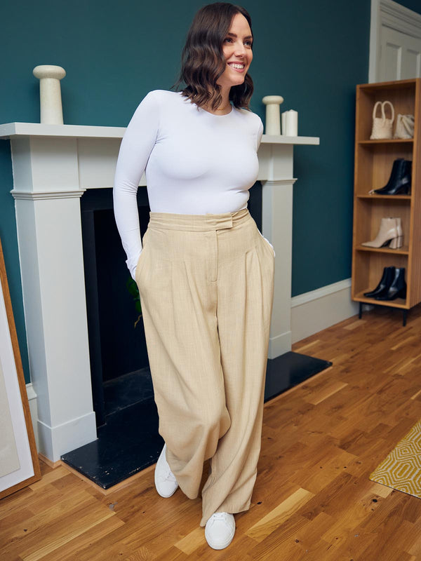 A model wearing the Lana trousers in sand and a long sleeve white top, pictured in front on a fireplace with their hands in their pockets. 