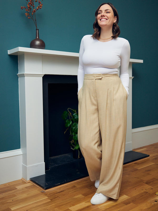 A model wearing the Lana trousers in sand and a long sleeve white top, pictured in front on a fireplace with their hands in their pockets.