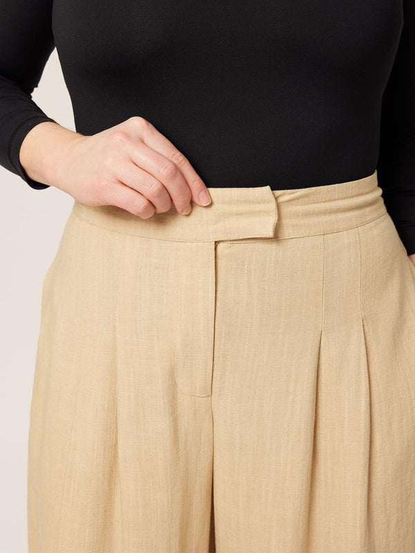 A close up of a model wearing the Lana trousers in sand, with a focus on the zip and clasp fastening.