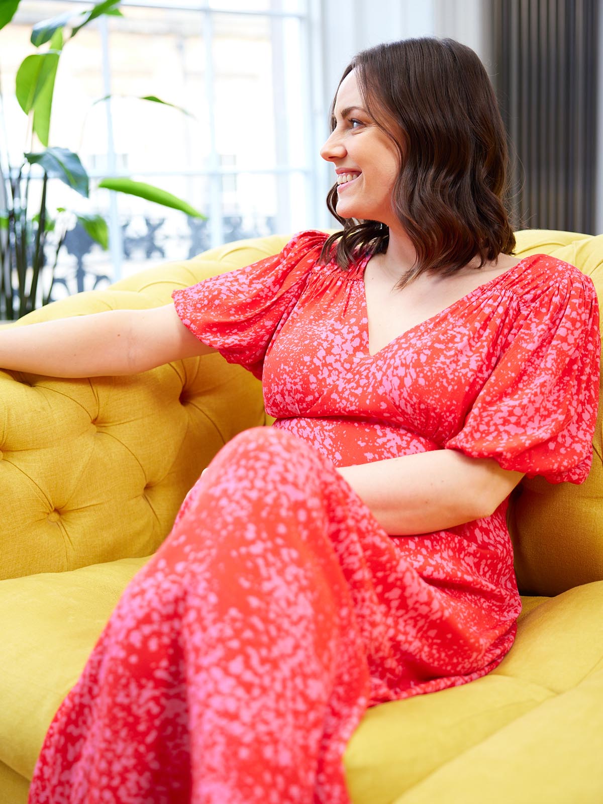 A model wears the Lauren midi dress in red and pink smudge print, pictured sitting on a bright yellow sofa in a living room.