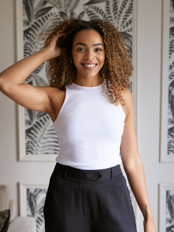 A model is pictured wearing the sustainable Abbey rib vest in white, holding one hand up to their hair and smiling into the camera. 