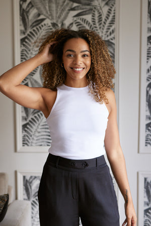 A model is pictured wearing the sustainable Abbey rib vest in white, holding one hand up to their hair and smiling into the camera. 