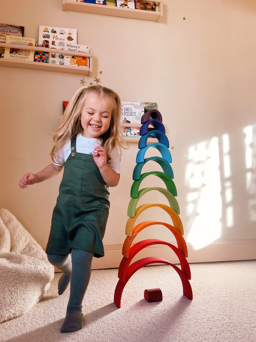 Picture of a smiling child wearing the sustainable green dungaree dress from This is Unfolded, playing in a playroom next to a rainbow stack