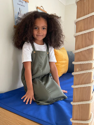 A child playing on a blue mat, pictured wearing the sustainable Lilah pinafore dress for kids. 