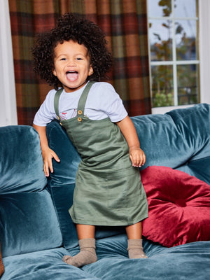 A child wearing the Lilah sustainable kids pinafore dress with pockets, pictured stood on a couch with a red velvet cushion.