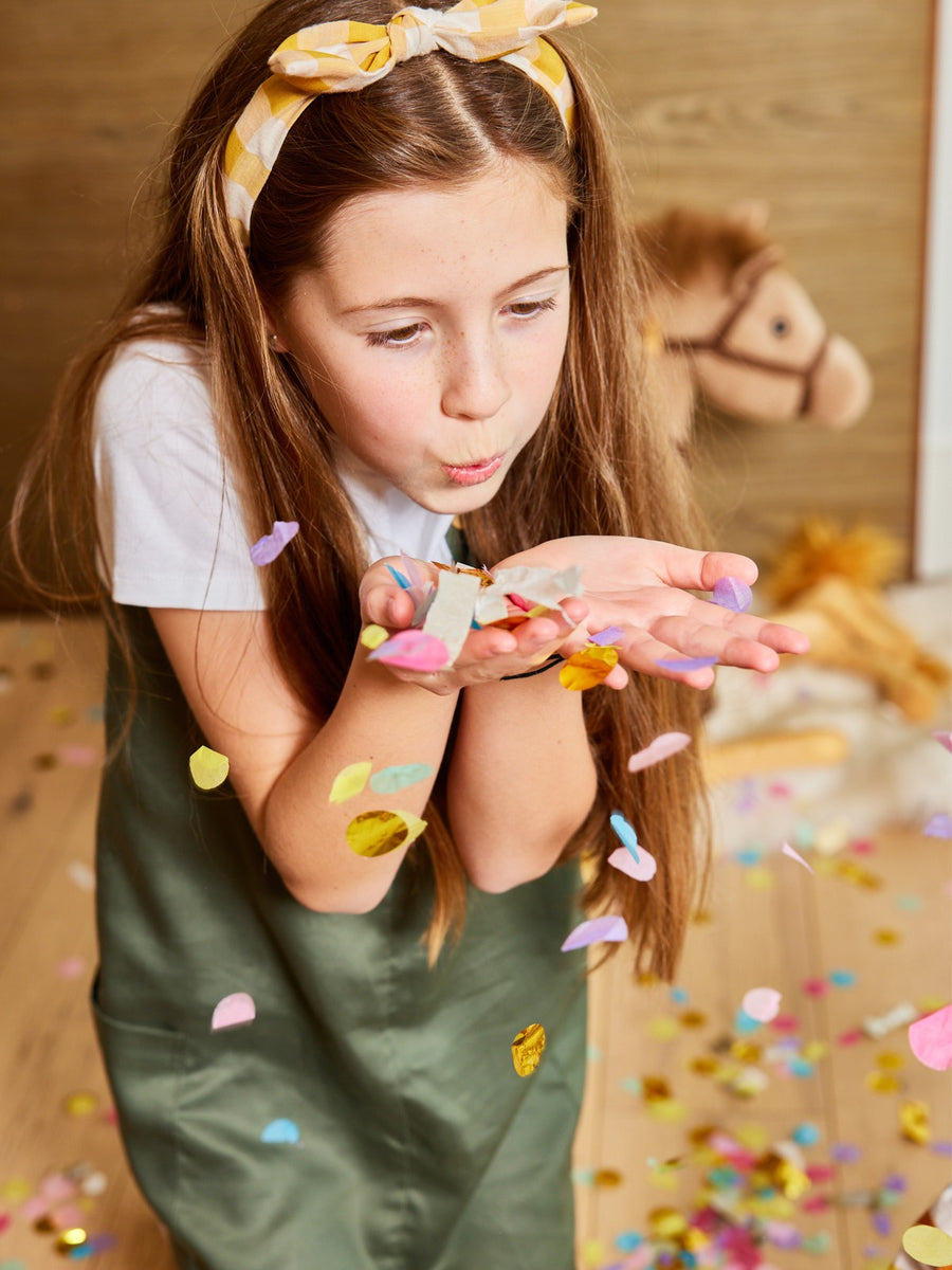 A child wearing the Lilah sustainable kids pinafore dress with pockets, pictured blowing confetti out of their hands. 
