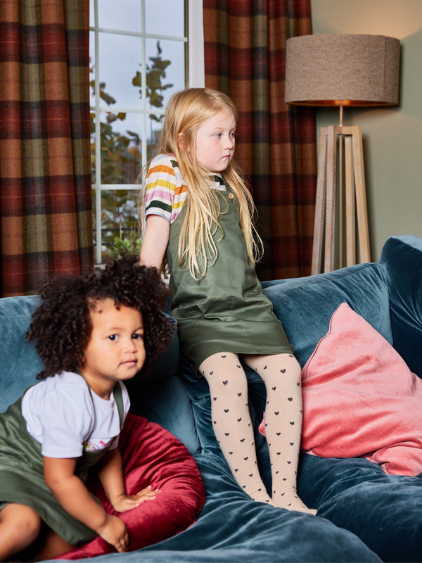 Two children wearing the Lilah sustainable kids pinafore dress with pockets, pictured on a couch in a cosy living room. 