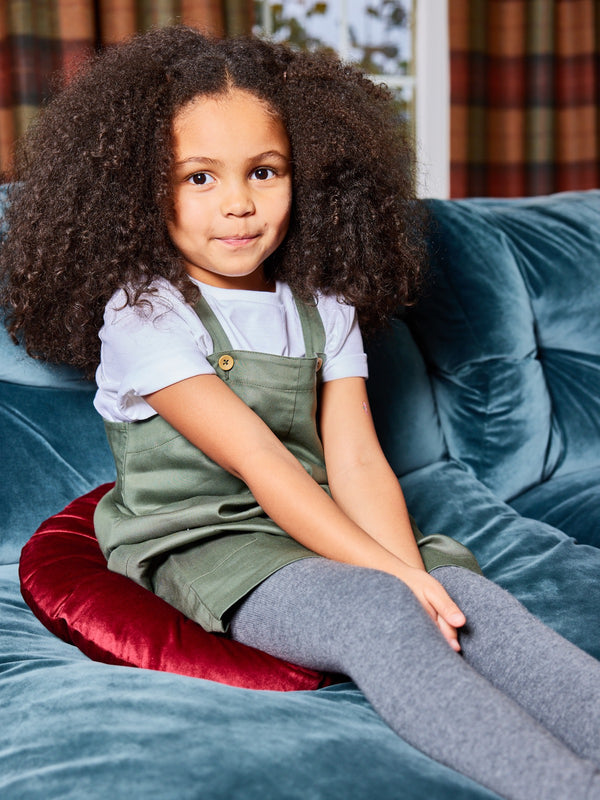 A child wearing the Lilah sustainable kids pinafore dress with pockets, pictured sat on a red cushion on a couch and smiling at the camera. 