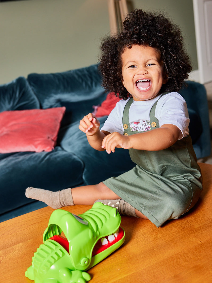 A happy child smiling widely, pictured with a toy crocodile and wearing the Lilah kids pinafore dress with pockets from sustainable clothing brand This is Unfolded. 