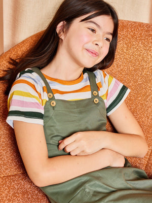 A child wearing the Lilah sustainable kids pinafore dress with pockets over the Hedy striped t-shirt, pictured sat on a couch with arms slightly crossed and smiling.