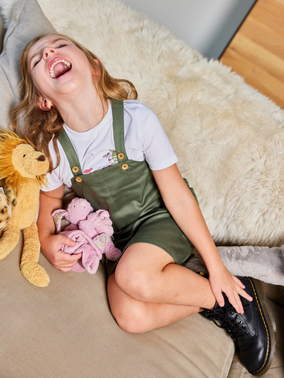 A child pictured laughing on a couch with cuddly toys, wearing the Lilah eco-friendly kids pinafore dress with pockets from This is Unfolded. 