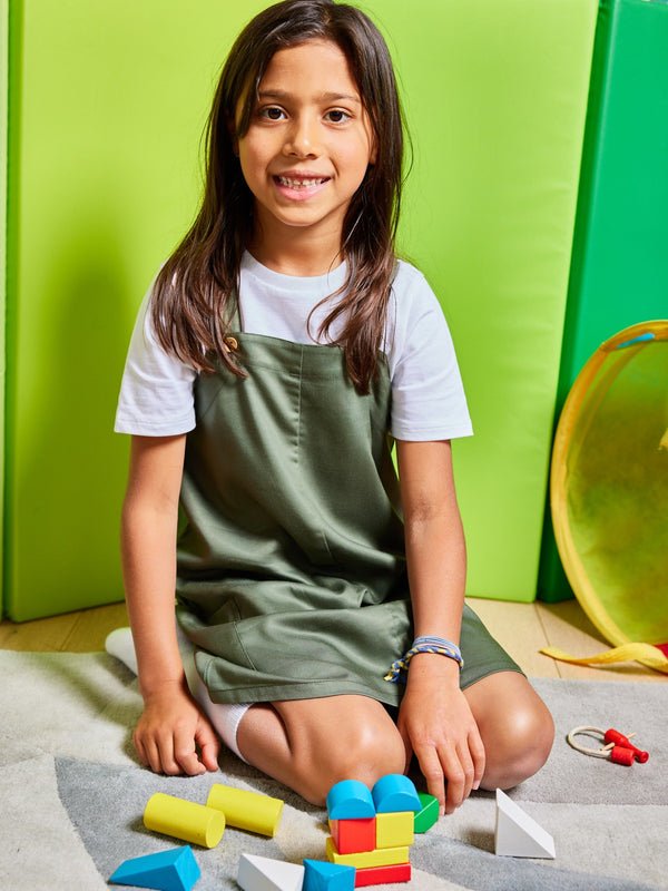 A child wearing the Lilah sustainable kids pinafore dress with pockets, pictured kneeling on a rug and playing with bright wooden toy blocks. 