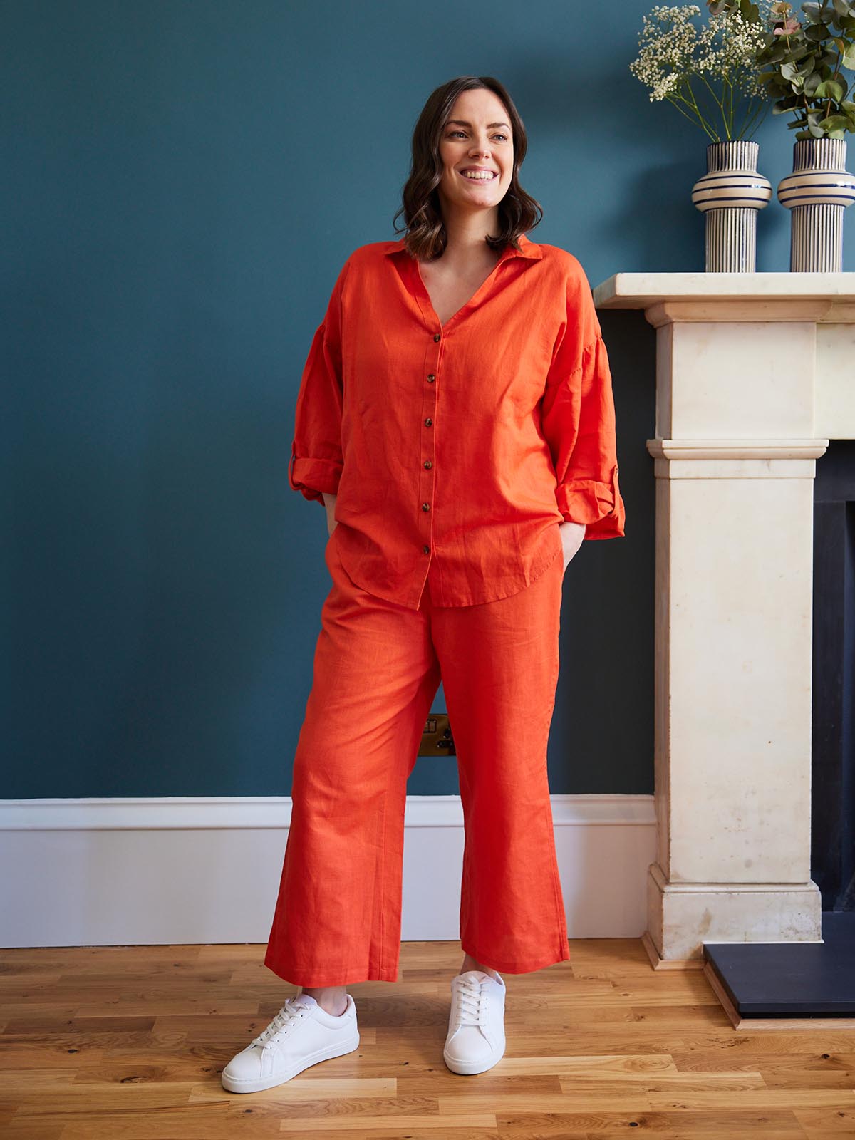 A model wears the Martha shirt and the Gigi trousers, both in a vibrant red, as a co-ordinating set. She's stood in a living room next to a fireplace with flowers on the mantlepiece. 