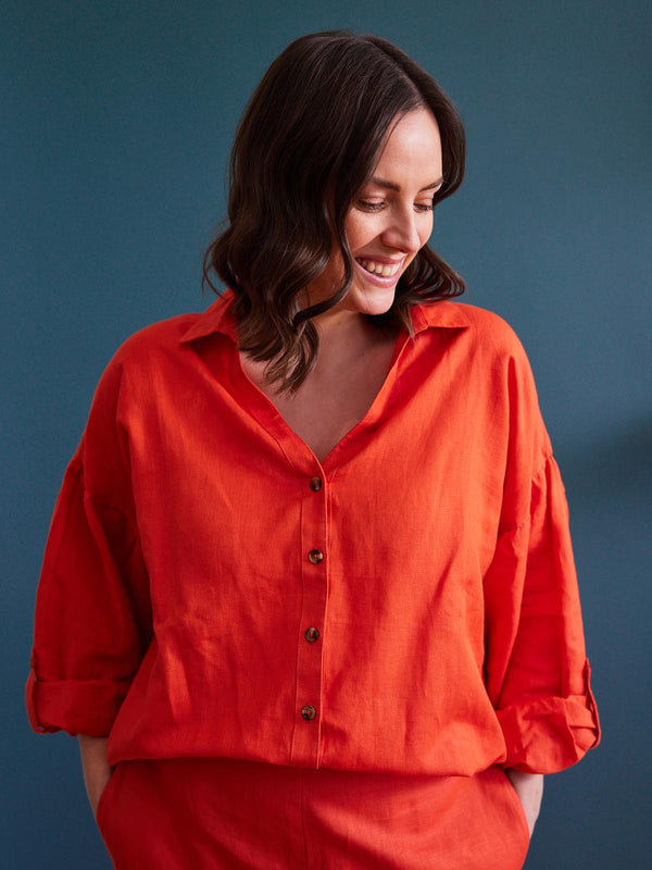 A model wears the Martha shirt and the Gigi trousers, both in a vibrant orange-red, as a co-ordinating set, pictured smiling with their hands in their pockets.