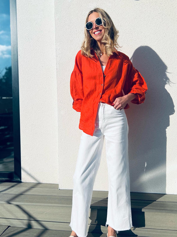 A woman wears the sustainable Martha shirt in red, tucked into a pair of white trousers. She is also wearing sunglasses and is stood outside on a sunny deck area. 
