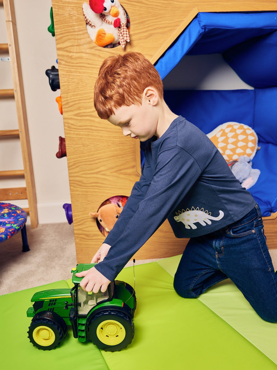 Child pictured playing with a green tractor toy on a green playroom mat, wearing the Melodi long sleeved kids t-shirt in navy blue with a dinosaur print from sustainable fashion brand This is Unfolded. 