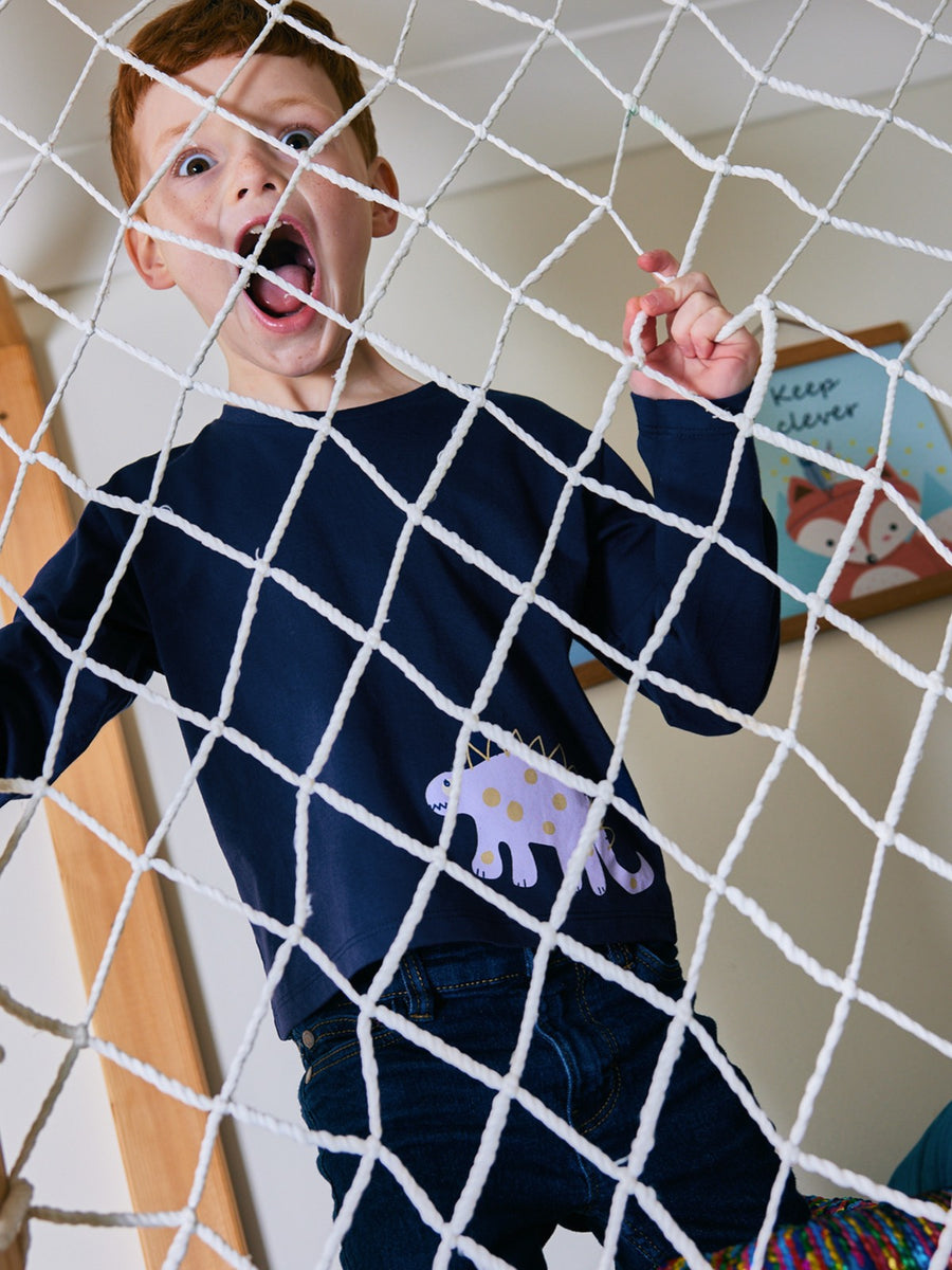 Child wearing the Melodi sustainable long sleeved kids t-shirt in navy blue with a dinosaur print, pictured holding on to the net of an indoor climbing frame in a playroom and pulling a playful and scary face. 