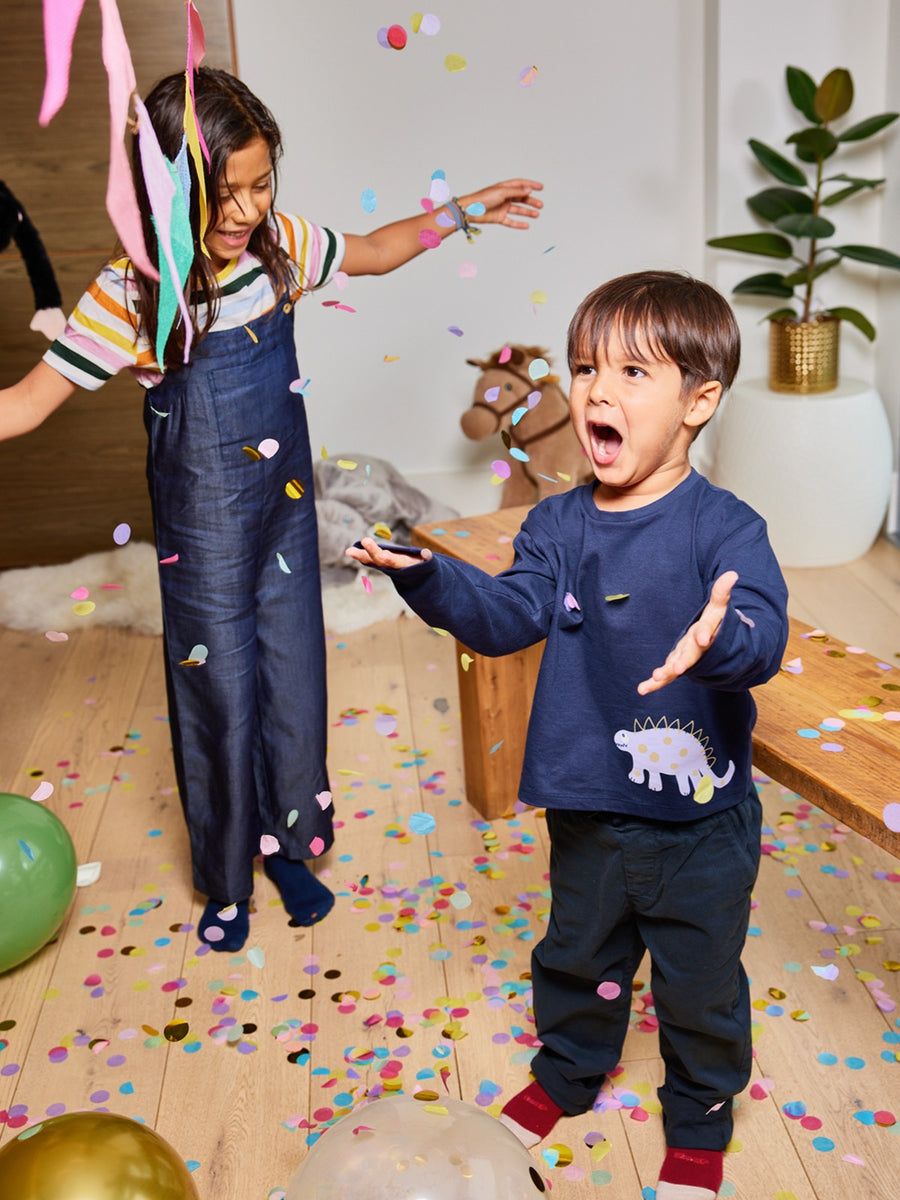 Two children pictured enjoying falling confetti in a party atmosphere, one wearing the Melodi sustainable long sleeved kids t-shirt in navy blue with a dinosaur print.