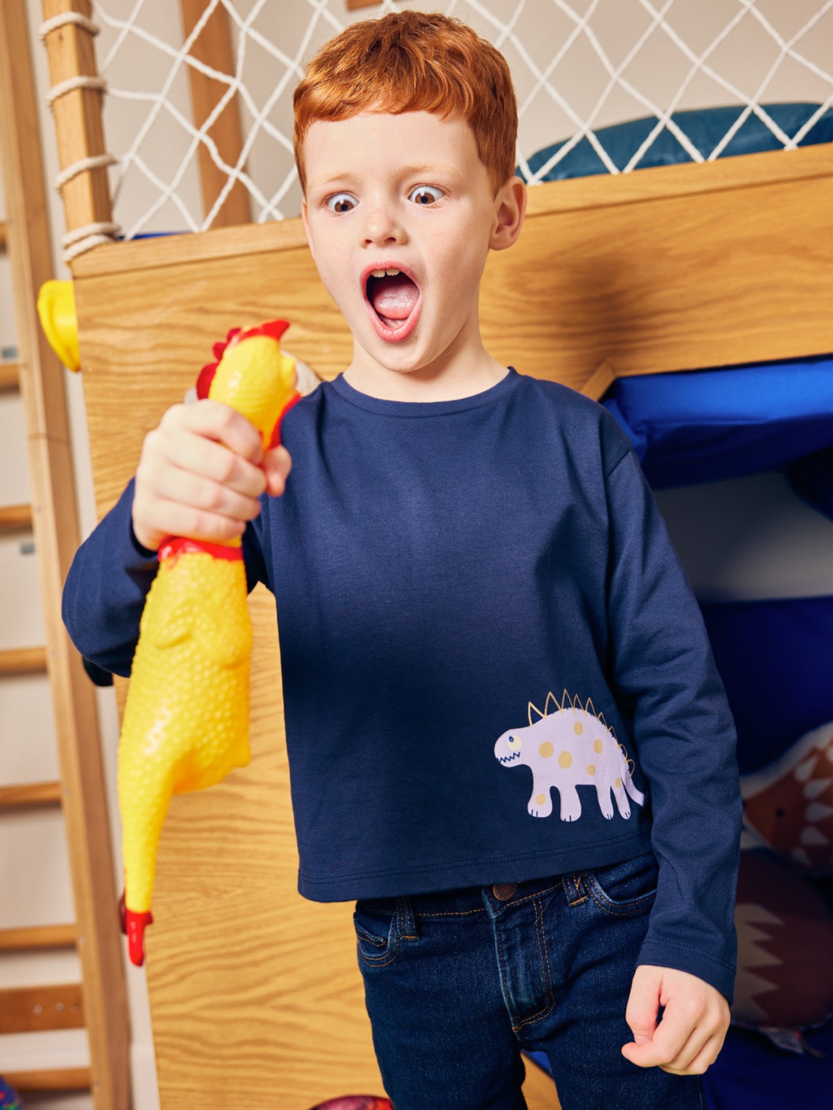 Child wearing the Melodi sustainable long sleeved kids t-shirt in navy blue with a dinosaur print, pictured staring comically wide mouthed at a chicken toy that they are holding. 