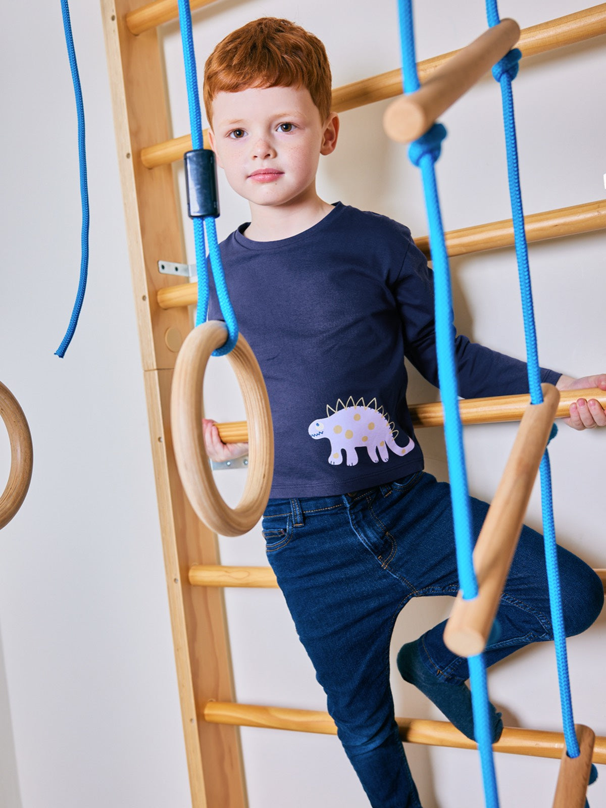 Child pictured on an indoor climbing frame, wearing the Melodi eco-friendly long sleeved kids t-shirt in navy blue with a dinosaur print from This is Unfolded. 
