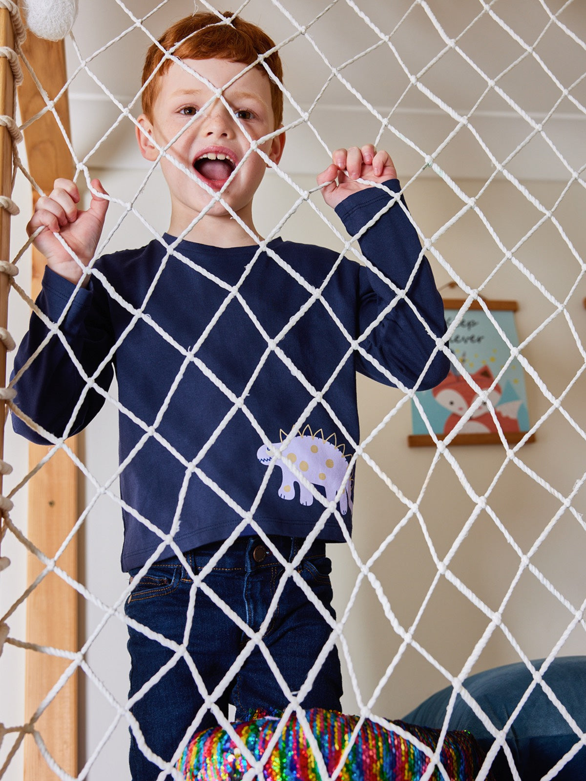 Child wearing the Melodi sustainable long sleeved kids t-shirt in navy blue with a dinosaur print, pictured holding on to the net of an indoor climbing frame in a playroom. 