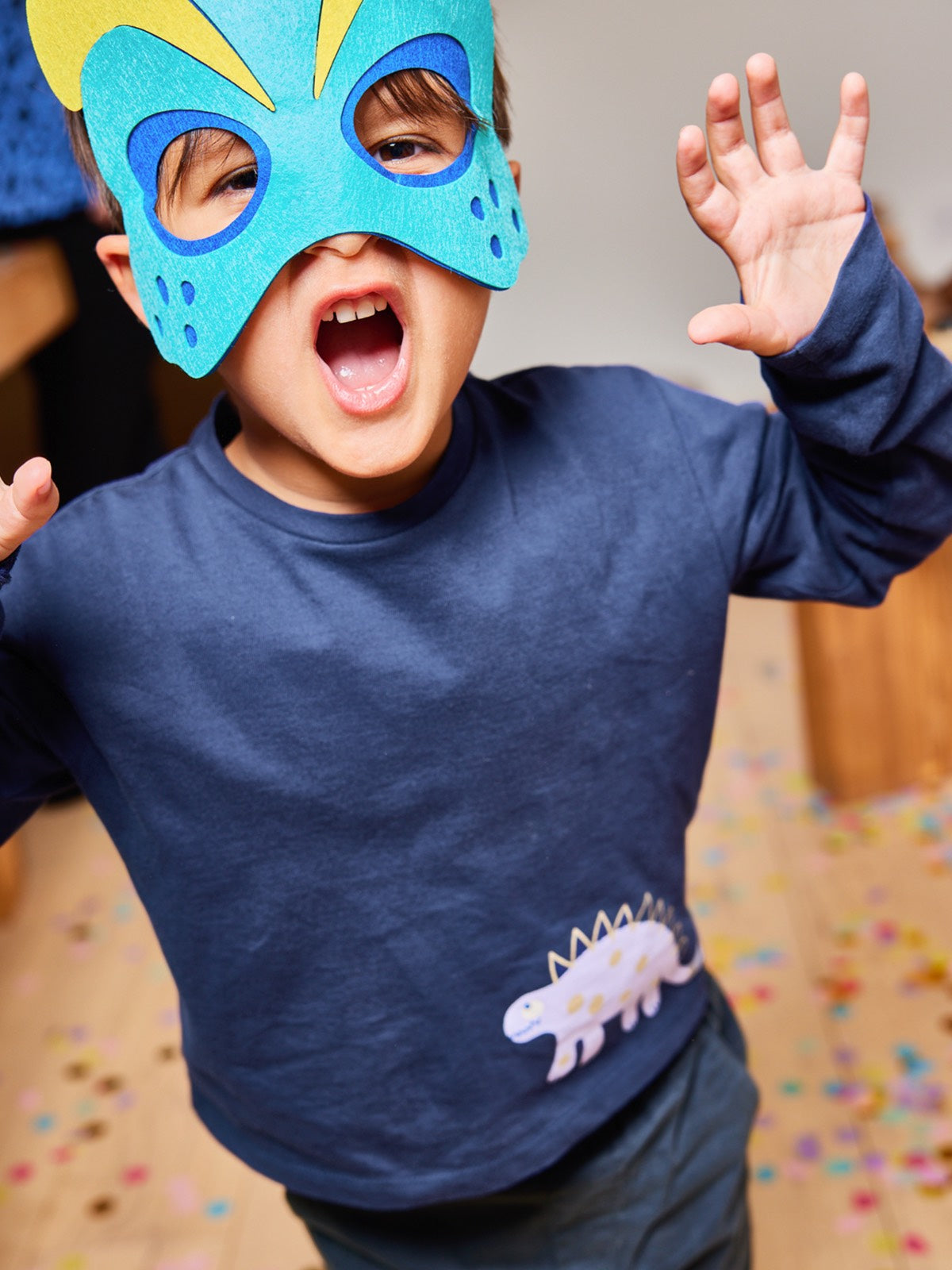 Child wearing the Melodi sustainable long sleeved kids t-shirt in navy blue with a dinosaur print, pictured wearing a colourful mask and imitating roaring at the camera. 