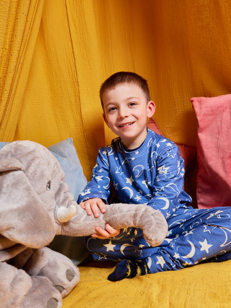A child wearing the Mira sustainable kids PJ set in blue with a white moon and star print, pictured sitting cross-legged on the floor with bright cushions, holding on to a cuddly toy and looking happy.