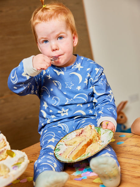 A child wearing the Mira sustainable kids PJ set in blue with a white moon and star print, pictured eating a slice of cake from a paper plate with a fork and looking into the camera. 