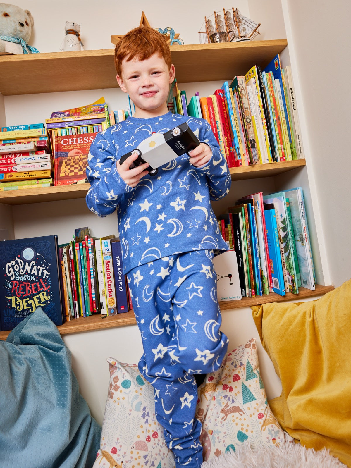 A child wearing the Mira sustainable kids PJ set in blue with a celestial print, pictured standing in front of a bookcase holding a book and smiling at the camera. 