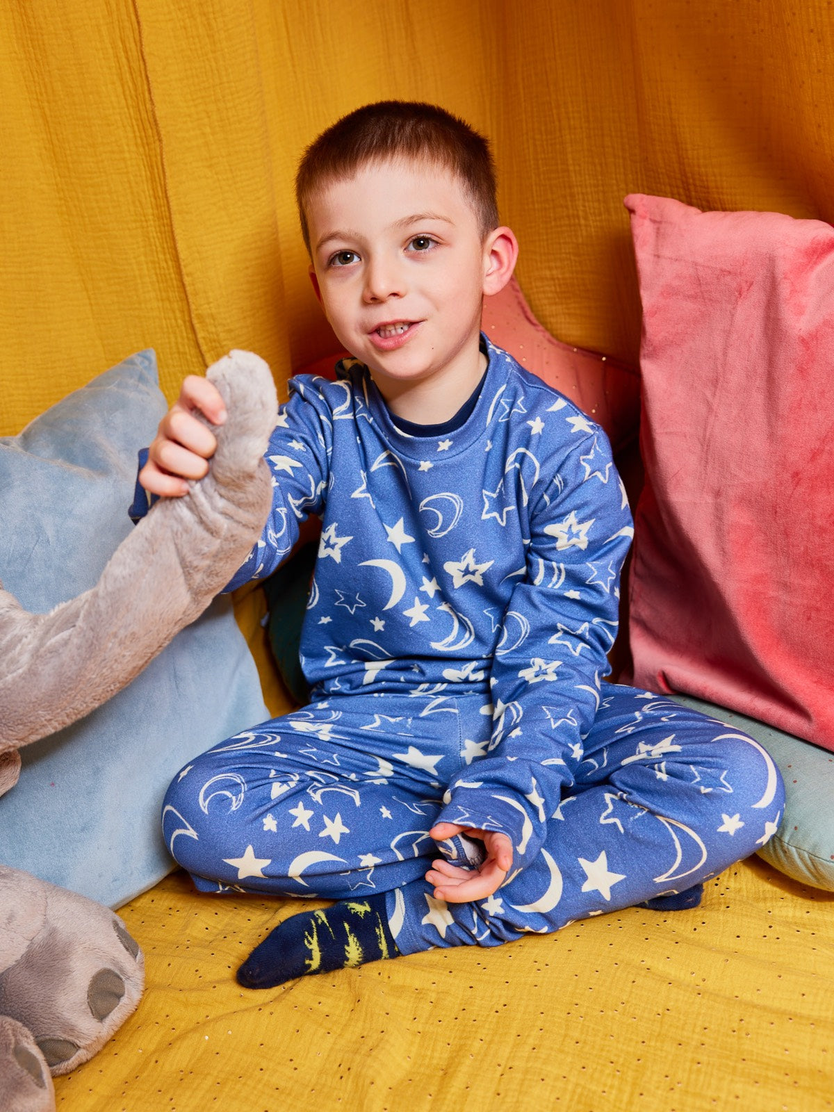 A child wearing the Mira sustainable kids PJ set in blue with a white moon and star print, pictured sitting cross-legged on the floor with bright cushions and holding on to a cuddly toy.