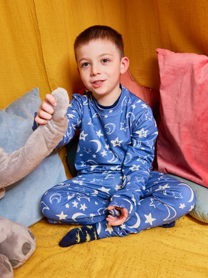 A child wearing the Mira sustainable kids PJ set in blue with a white moon and star print, pictured sitting cross-legged on the floor with bright cushions and holding on to a cuddly toy.