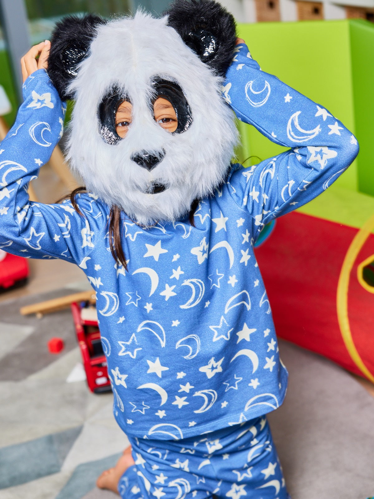 A child wearing the Mira sustainable kids PJ set in blue with a white moon and star print, pictured in a playroom wearing a fluffy panda mask. 