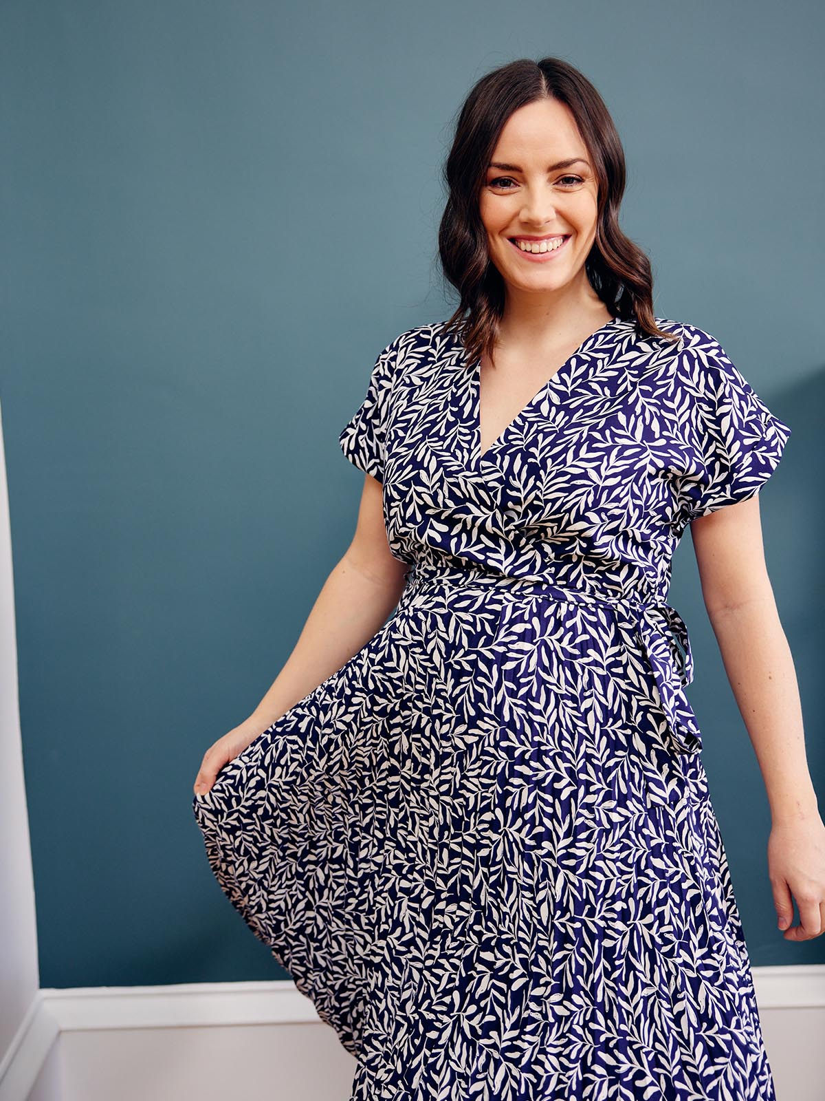 A model wears the Nena pleated wrap dress in navy abstract leaf print, pictured smiling at the camera and holding up one side of the dress slightly. 