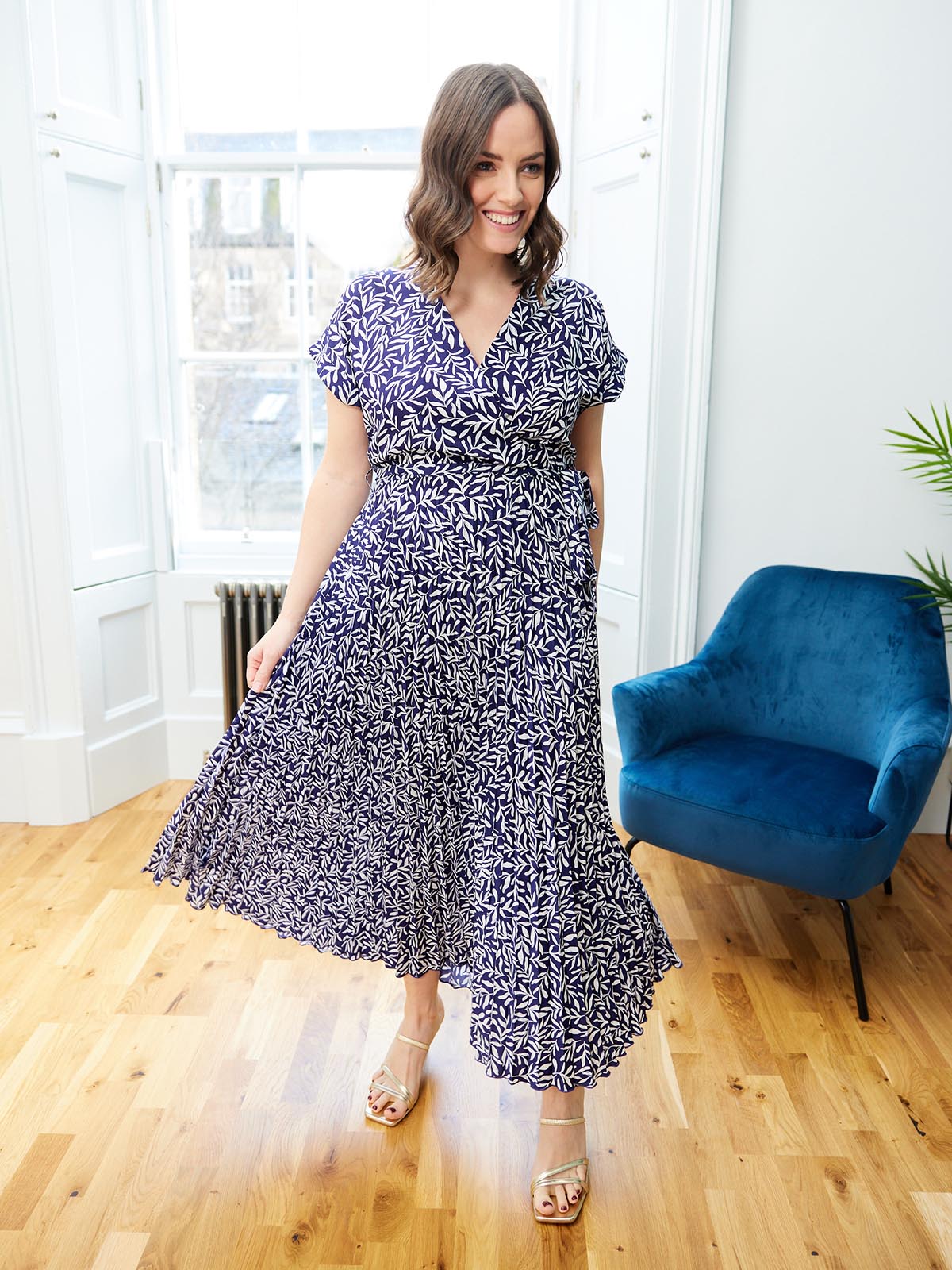 A model stands in a well lit room, wearing the sustainable Nena dress from This is Unfolded. They are holding the pleated fabric out slightly and smiling. 