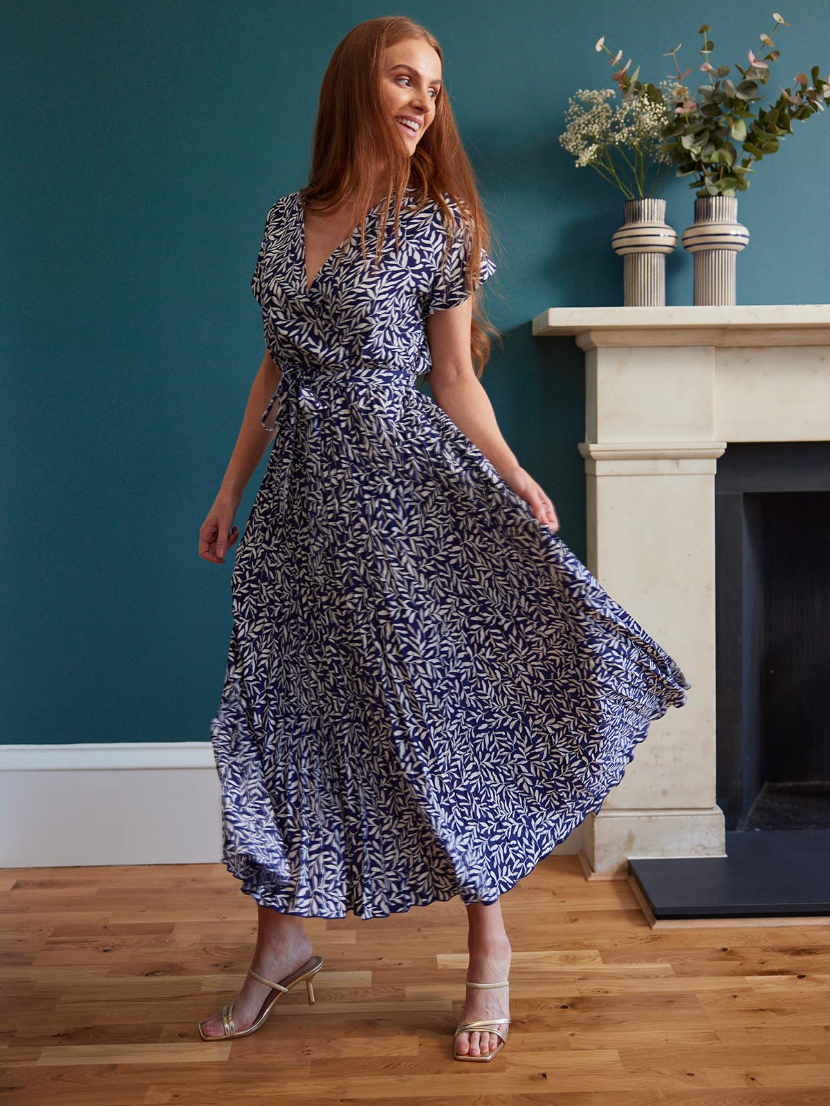 A model stands in front of a fireplace in a living area, wearing the sustainable Nena dress from This is Unfolded. They are holding the pleated fabric out slightly and smiling.