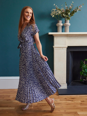 A model stands in front of a fireplace in a living area, wearing the sustainable Nena dress from This is Unfolded. They are holding the pleated fabric out slightly and smiling.