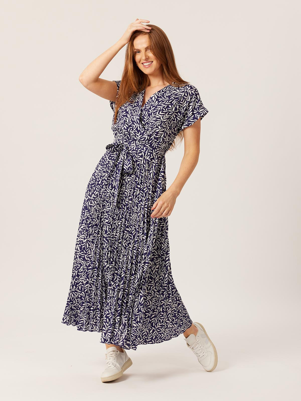 A model wears the Nena Wrap Dress from Unfolded, in navy abstract leaf print. They are pictured running their hand through their hair and smiling into the camera. 