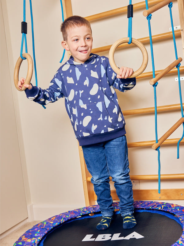 A child wearing the Seb sustainable kids hoodie in navy with a blue and white terrazzo print, pictured on a mini trampoline in front of an indoor climbing area.