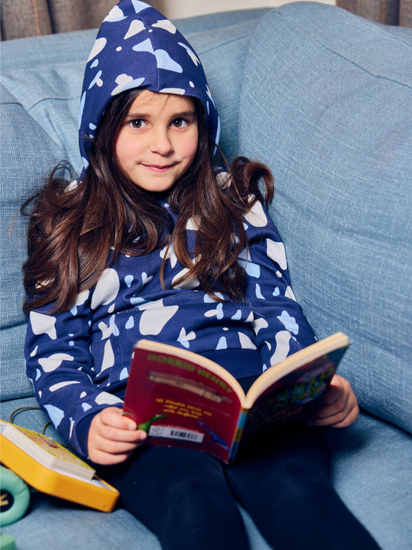 A child wearing the Seb sustainable kids hoodie in navy with a blue and white terrazzo print, pictured on a couch reading a book with their hood up and looking cosy. 