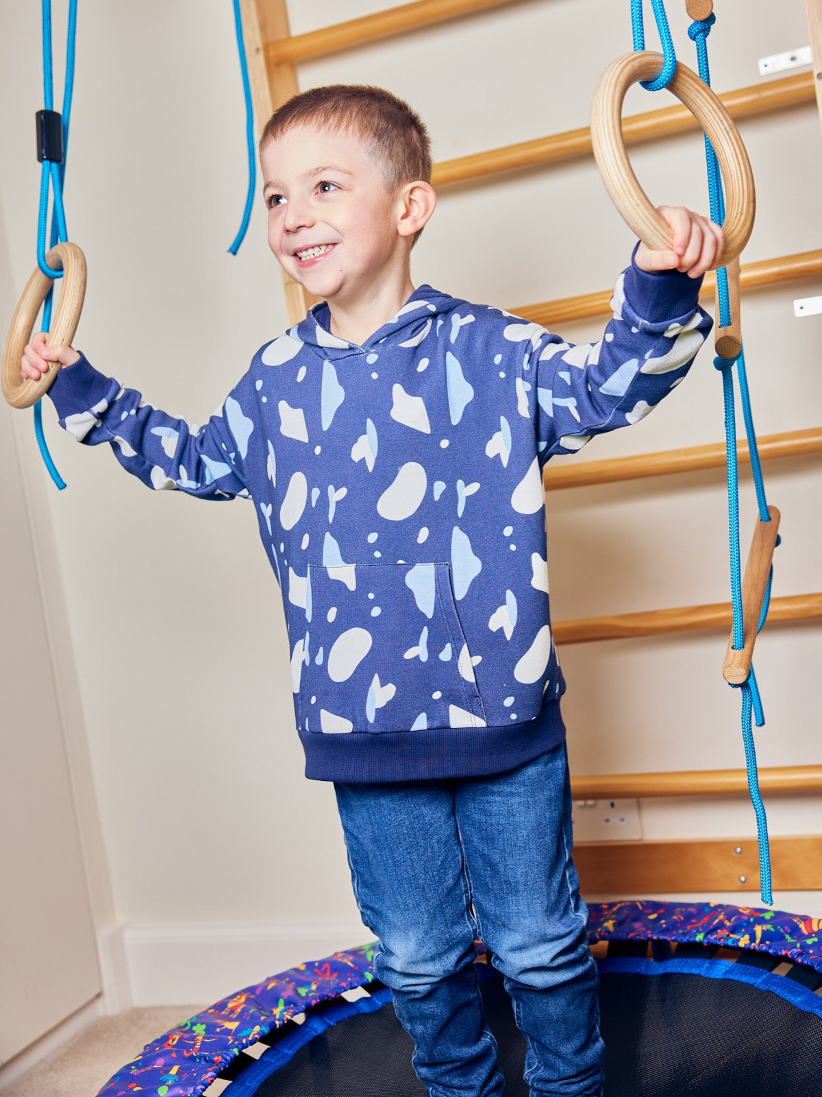 A child wearing the Seb sustainable kids hoodie in navy with a blue and white terrazzo print, pictured standing on a mini trampoline and holding on to an indoor climbing frame. 