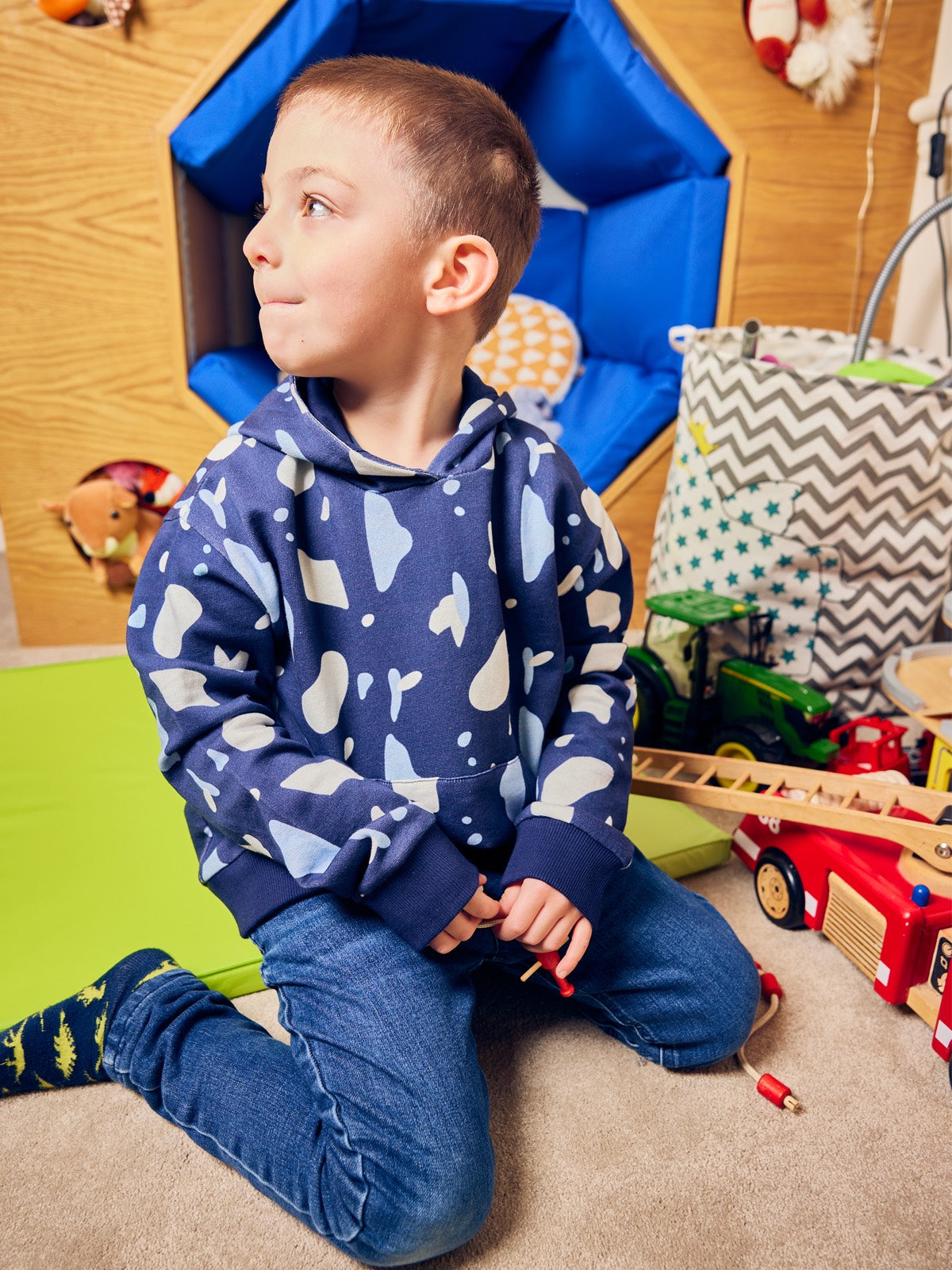 A child wearing the Seb sustainable kids hoodie in navy with a blue and white terrazzo print, pictured on the floor of a playroom and playing with colourful toys. 