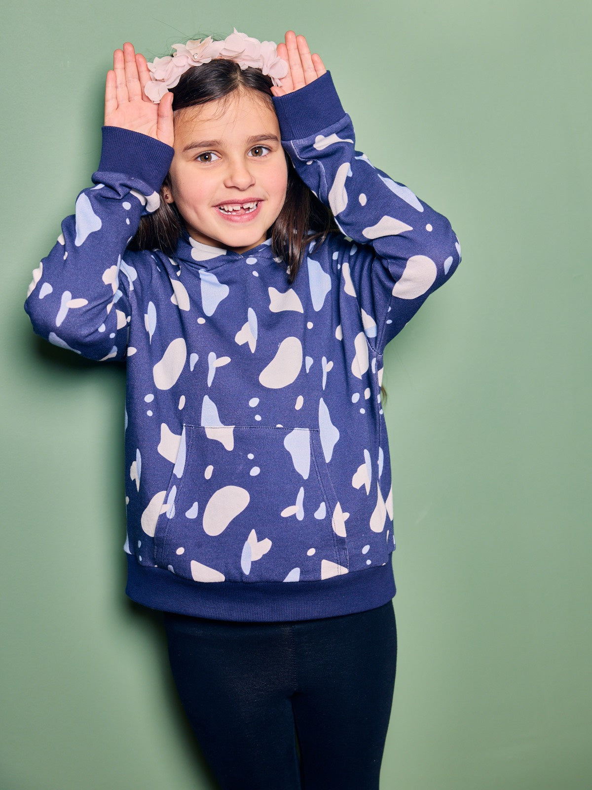 A child wearing the Seb sustainable kids hoodie in navy with a blue and white terrazzo print, pictured with their hands by their head like bunny ears and looking into the camera. 